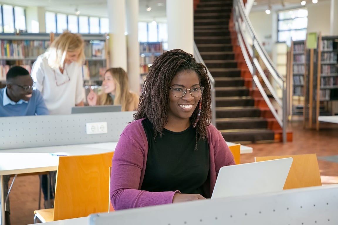 Un étudiant afro-américain navigue sur un ordinateur portable alors qu'il est assis à une table dans une bibliothèque avec des camarades de classe et un professeur.